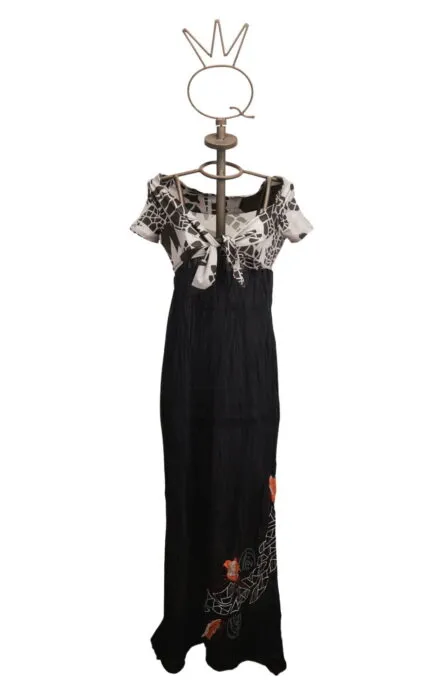 Abito lungo da donna con maniche corte e scollatura a V, caratterizzato da un top in bianco e nero con motivo floreale e una gonna nera con ricami colorati. Esposto su un manichino con una base decorativa.