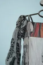 Blusa elegante in tessuto leggero con maniche ampie, caratterizzata da un mix di colori e motivi artistici, esposta su un manichino. Sfondo azzurro.