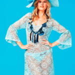 Model in transparent dress with circle pattern and bell sleeves, wears a light blue hat decorated with flowers, on a blue background. Ideal for summer and beach looks. 