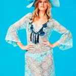 Modella in abito trasparente con motivo a cerchi e maniche a campana, indossa un cappello azzurro decorato con fiori, su sfondo blu. Ideale per look estivi e da spiaggia.