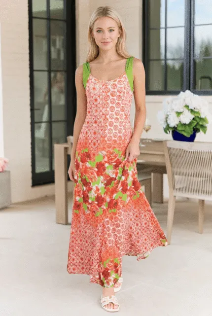 Modella che indossa un abito lungo estivo con stampa floreale in tonalità arancione e verde, posizionata in un ambiente luminoso e moderno con piante e arredi eleganti.