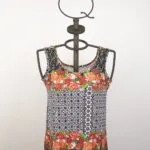 Top estivo a maniche corte con motivo floreale e geometrico, presentato su un manichino in metallo. Design vivace con fiori rossi e dettagli in bianco e nero, ideale per outfit casual e freschi.