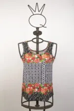 Top estivo a maniche corte con motivo floreale e geometrico, presentato su un manichino in metallo. Design vivace con fiori rossi e dettagli in bianco e nero, ideale per outfit casual e freschi.