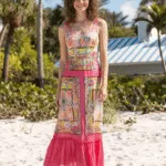 Modella che indossa un abito lungo estivo dai colori vivaci, con dettagli floreali e una gonna in chiffon, posando sulla spiaggia con palme sullo sfondo.