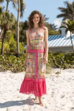 Modella che indossa un abito lungo estivo dai colori vivaci, con dettagli floreali e una gonna in chiffon, posando sulla spiaggia con palme sullo sfondo.