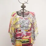 Maglietta colorata a maniche larghe esposta su un manichino, caratterizzata da motivi floreali e strisce vivaci, ideale per un look estivo e casual.