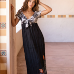 Modella in abito lungo nero con dettagli floreali, posa in un ambiente luminoso con pareti beige e decorazioni in ceramica. I suoi capelli ondulati e il trucco naturale mettono in risalto la bellezza del look estivo.
