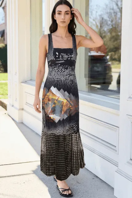 Modella che indossa un abito lungo a spalle scoperte con un design grafico moderno, in piedi davanti a una finestra. L'abito presenta una combinazione di colori scuri con un motivo artistico vivace al centro, perfetto per eventi estivi o occasioni casual.