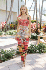 Modella che indossa un abito lungo e colorato con dettagli floreali, mentre posa in un ambiente all'aperto con piante e alberi. L'abito presenta una parte superiore in pizzo rosso e un design vivace.