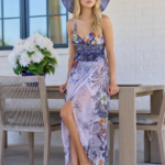 Modella con abito estivo floreale e cappello di paglia, seduta a un tavolo all'aperto, circondata da fiori bianchi in un vaso, in un ambiente luminoso e moderno.