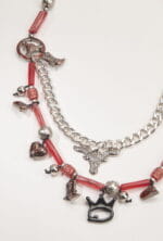 Collana decorativa con elementi in metallo e perline rosse, caratterizzata da charms a forma di stivali, cuore e teschio, con una catena argentata e un ciondolo a forma di testa di toro.