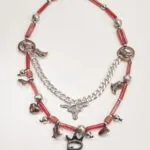 Collana artistica con perline rosse e pendenti metallici, tra cui stivali, cuori e un simbolo di toro, perfetta per un look originale e creativo.