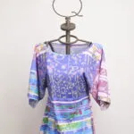 Maglietta colorata con maniche a 3/4 esposta su un manichino, caratterizzata da un design vivace e grafico, ideale per la moda estiva e casual.