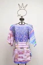 Blusa colorata con maniche a sbuffo, presentata su un manichino, caratterizzata da un mix di motivi geometrici e floreali in tonalità di blu, viola e rosa. Ideale per un look estivo e vivace.
