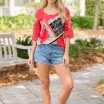 T-shirt lunga in tessuto stretch di colore rosso con grafica moderna, indossata da una giovane donna in un ambiente all'aperto, abbinata a shorts in denim e sandali.