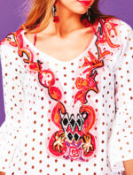 Tunica bianca con ricami colorati e dettagli floreali, indossata da una giovane donna con orecchini rossi, su sfondo viola. Perfetta per look estivi e occasioni informali.