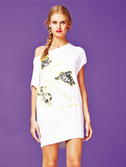 Modella che indossa una tunica bianca con stampa di tartarughe e dettagli colorati su sfondo viola. La tunica ha un design asimmetrico e maniche corte, ideale per un look estivo e casual.
