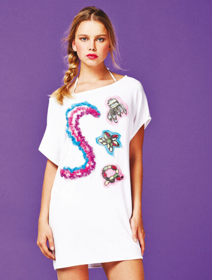 Tunica bianca con ricami colorati e dettagli decorativi, indossata da una giovane donna su sfondo viola. Perfetta per look estivi e casual.