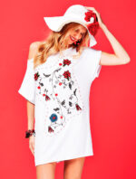 Modella che indossa una tunica bianca con ricami floreali colorati, abbinata a un cappello bianco e un bracciale. Sullo sfondo, un vivace colore rosso che mette in risalto l'outfit estivo.