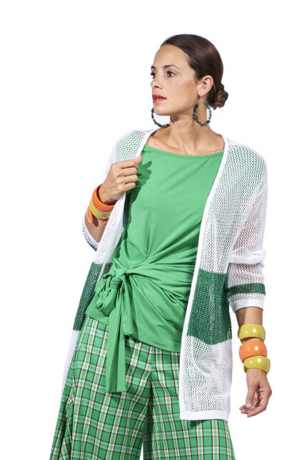 Donna elegante che indossa un cardigan bianco e verde, abbinato a una maglietta verde e pantaloni a quadri. Accessori colorati completano il look moderno e fresco.