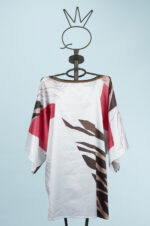 Blusa stampata con maniche ampie, caratterizzata da un design moderno con motivi astratti in tonalità di rosso e marrone, esposta su un manichino su sfondo azzurro.