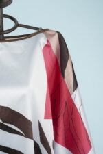 Blusa stampata con motivi grafici in tonalità rosse, marroni e beige, appesa su un manichino su sfondo azzurro.