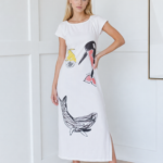 Modella che indossa un vestito lungo bianco con stampe artistiche di pesci e uccelli, abbinato a sandali bianchi, in un ambiente moderno e luminoso.