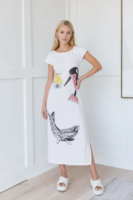 Modella che indossa un vestito lungo bianco con stampe artistiche di pesci e uccelli, abbinato a sandali bianchi, in un ambiente moderno e luminoso.